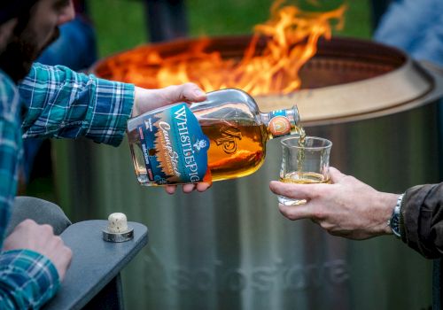 Two people by a fire pit: one pours a bottle labeled 